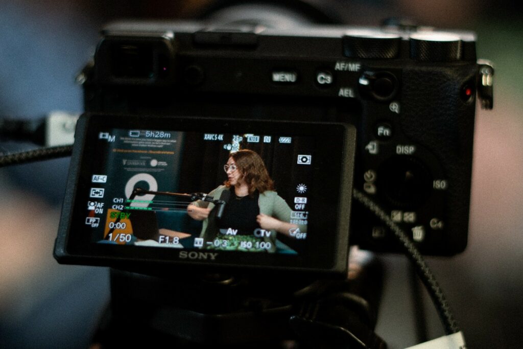 A close up photo of a camera preview screen which shows a woman speaking into a microphone on the set of a podcast